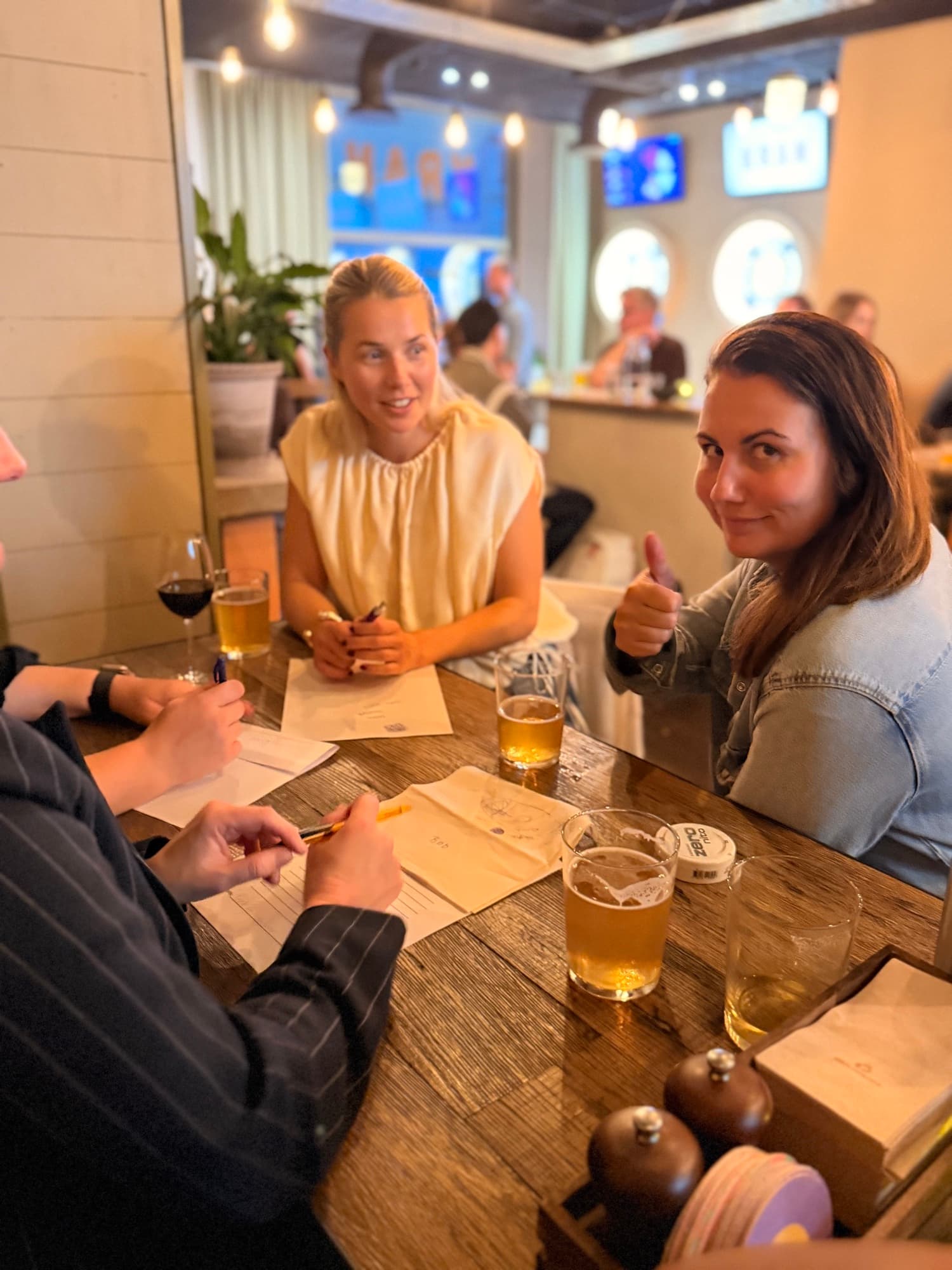 Quizlag runt ett bord med svarslappar och öl, en av gästerna ger tummen upp